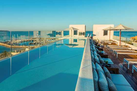 Näköala infinity-altaalle, jossa on aurinkotuoleja ja varjoja, ympärillä lasikaiteet ja taustalla satama. (Tekoälyn luoma teksti)