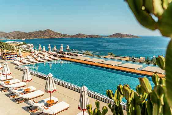 Uima-allas, jossa aurinkotuoleja ja päivänvarjoja, sijaitsee rannikon äärellä, näkymänä sininen meri ja vuoret. (Tekoälyn luoma teksti)