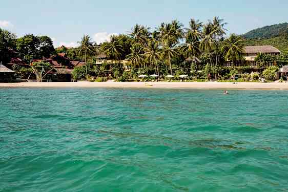 Näkymä hiekkarannalle, jota ympäröivät trooppiset puut ja rakennukset, vesiltä katsottuna. (Tekoälyn luoma teksti)