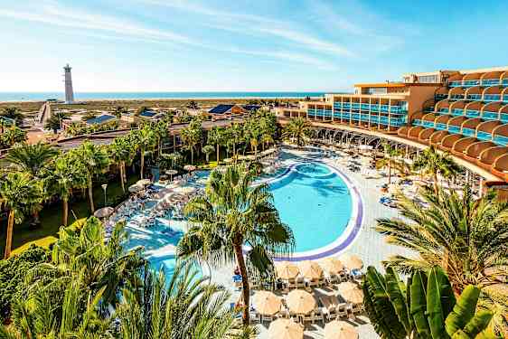 Hotellialue, jossa suuri uima-allas, aurinkotuoleja ja päivänvarjoja palmujen ympäröimänä, taustalla meri ja majakka. (Tekoälyn luoma teksti)