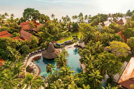 Ilmakuva trooppisesta lomakohteesta, jossa on uima-allasalue, vehreää kasvillisuutta ja perinteisesti rakennettuja bungaloweja. (Tekoälyn luoma teksti)