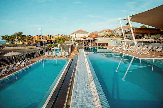 Aurinkoinen lomakohde, jossa kaksi ulkouima-allasta ja ympärillä aurinkotuoleja, taustalla rakennuksia. (Tekoälyn luoma teksti)