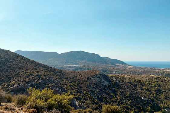 Maisema mäkisestä maastosta, jossa on vaihtelevaa kasvillisuutta, ja taustalla näkyy meri kirkkaan sinisen taivaan alla. (Tekoälyn luoma teksti)