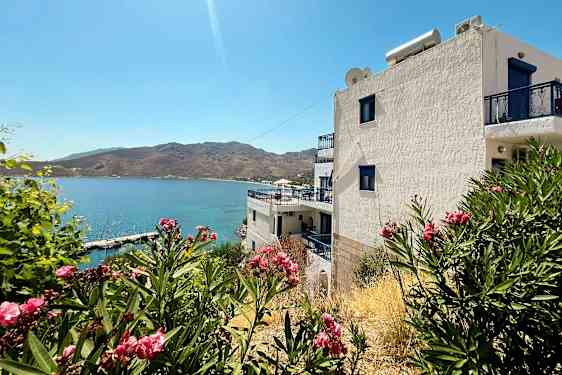 Valkoinen rakennus sinisillä yksityiskohdilla meren äärellä, ympärillä pensaita, joissa on vaaleanpunaisia kukkia. (Tekoälyn luoma teksti)