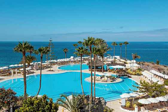 Näkymä suurelle uima-altaalle, jonka ympärillä on aurinkotuoleja ja aurinkovarjoja, sekä meri taustalla. (Tekoälyn luoma teksti)