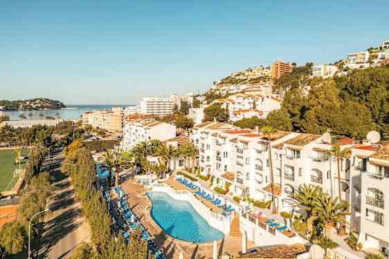 Kuva hotellialueesta, jossa on uima-allas ja aurinkotuoleja, ympäröivänä puita ja rakennuksia sekä näkymä merelle. (Tekoälyn luoma teksti)