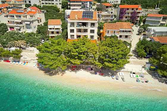 Ilmakuva rannasta, jossa on aurinkotuoleja, varjoja ja kirkasta vettä rakennusten edessä, joita ympäröivät puut. (Tekoälyn luoma teksti)