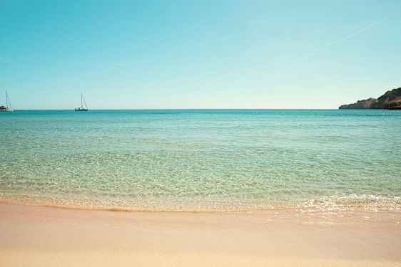 Ranta, jossa kirkasta vettä ja kaksi purjevenettä merellä. (Tekoälyn luoma teksti)