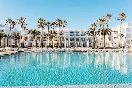 Hotellin allasalue, jossa kirkkaansinistä vettä, ympäröitynä aurinkotuoleilla, aurinkovarjoilla ja palmuilla. Taustalla näkyy valkoinen rakennus parvekkeineen. (Tekoälyn luoma teksti)