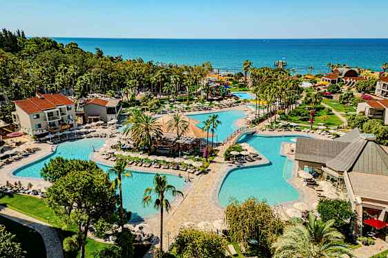 Lomakeskus, jossa on useita uima-altaita, ympäröitynä vehreydellä ja rakennuksilla, taustalla meri. (Tekoälyn luoma teksti)