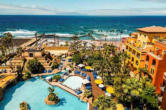 Hotellialue, jossa on uima-altaita, kasvillisuutta ja värikkäitä rakennuksia meren rannalla. (Tekoälyn luoma teksti)