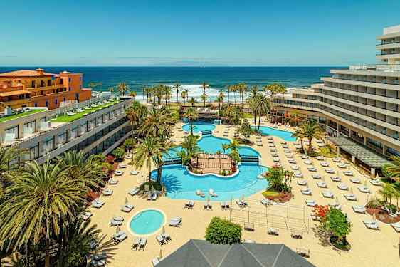 Hotellikompleksi, jossa on uima-altaita, aurinkotuoleja ja palmuja, lähellä merta. (Tekoälyn luoma teksti)