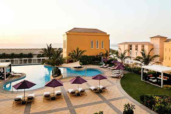 Ulkouima-allas, jonka ympärillä on aurinkotuoleja ja aurinkovarjoja, palmujen ja keltaisten rakennusten ympäröimänä. (Tekoälyn luoma teksti)