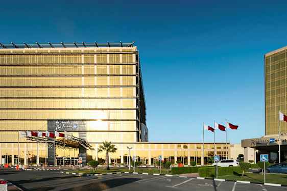 Moderni rakennus, jossa on kultaväriset lasiseinät, lipputankoja ja pysäköintialue. (Tekoälyn luoma teksti)