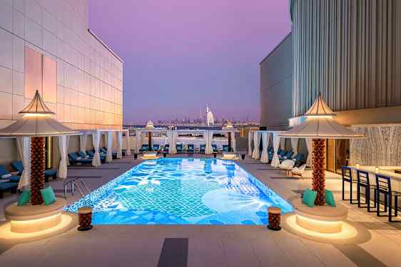Kattoterassi, jossa koristeellinen uima-allas, jota ympäröivät aurinkotuolit ja pienet aurinkovarjot. Taustalla näkyy Burj Al Arab ja Dubain kaupunkimaisema violetin iltataivaan alla. (Tekoälyn luoma teksti)