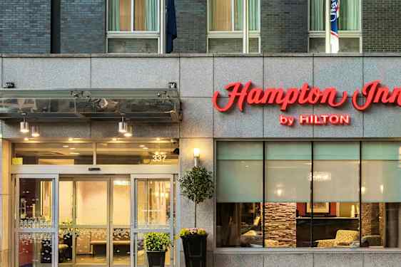 Hotellin sisäänkäynti, jonka yläpuolella lukee "Hampton Inn by Hilton". (Tekoälyn luoma teksti)