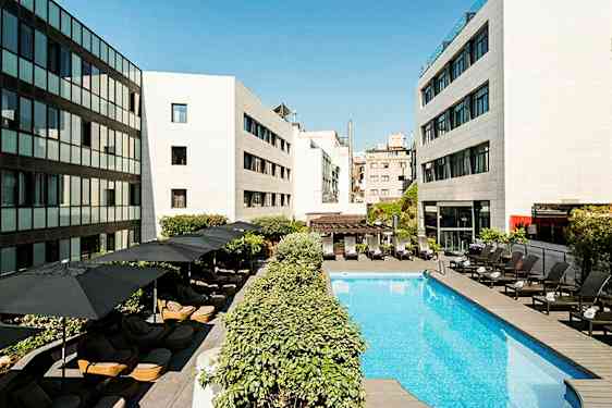 Hotellin uima-allas, jonka ympärillä on aurinkotuoleja ja varjoja, modernit rakennukset ympärillä. (Tekoälyn luoma teksti)