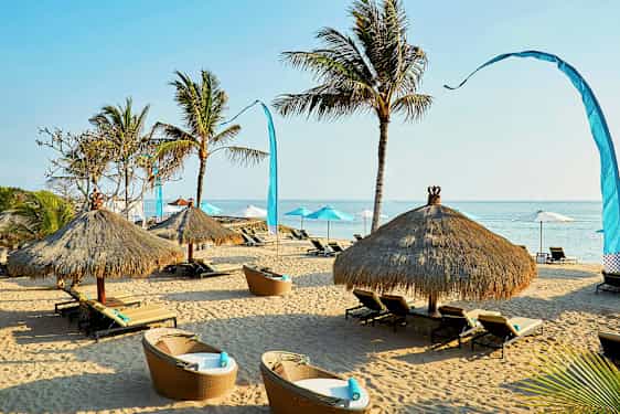 Hiekkaranta, jossa on aurinkotuoleja ja olkatattikatoksia, joita ympäröi muutama palmu. (Tekoälyn luoma teksti)