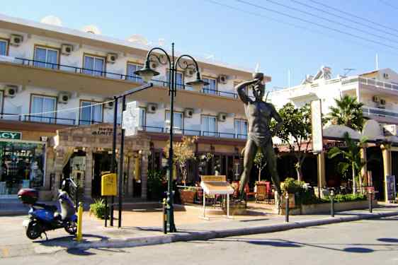 Patsas rakennuksen edessä, jossa on kauppoja ja ravintola. Skootteri on pysäköity vieressä. (Tekoälyn luoma teksti)