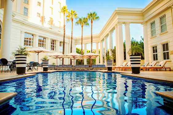 Uima-allasalue, jossa kirkasta sinistä vettä, aurinkotuoleja ja päivänvarjoja pylväiden ja palmujen ympäröimänä. (Tekoälyn luoma teksti)