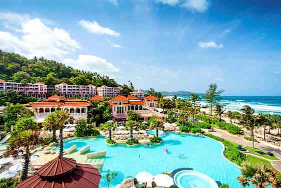 Kuva ylellisestä hotellialueesta meren rannalla, jossa on suuri uima-allas palmun ympäröimänä ja rakennuksia punaisilla katoilla. (Tekoälyn luoma teksti)