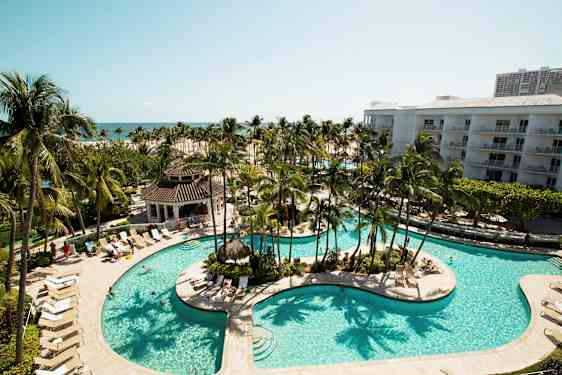 Kuvassa on ylellinen lomakeskus, jossa on suuri uima-allas ympärillään aurinkotuoleja ja palmuja. Taustalla näkyy lisää rakennuksia ja meri. (Tekoälyn luoma teksti)