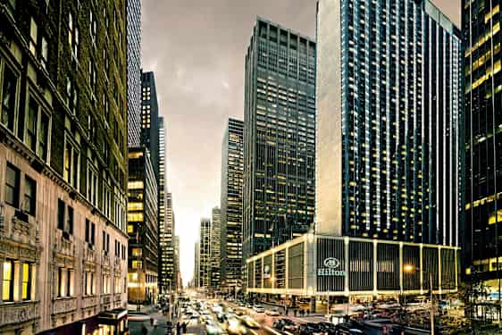 Kaupunki näkymä, jossa on korkeita pilvenpiirtäjiä, vilkasta liikennettä ja rakennuksia, joissa on lukuisia ikkunoita. Hilton-hotelli näkyy alareunassa. (Tekoälyn luoma teksti)