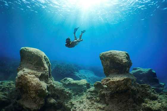 Snorklaaja ui veden alla suurten kallioiden välissä, kun auringonvalo suodattuu pinnalta. (Tekoälyn luoma teksti)