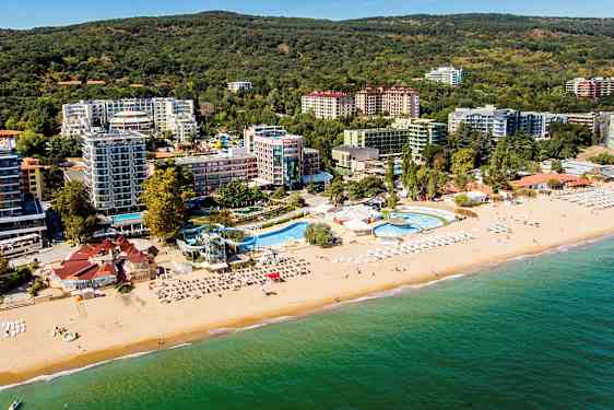 Ilmakuva rannasta, jossa on aurinkotuoleja ja aurinkovarjoja, uima-allasalue ja hotelleja viheralueiden ympäröimänä. (Tekoälyn luoma teksti)