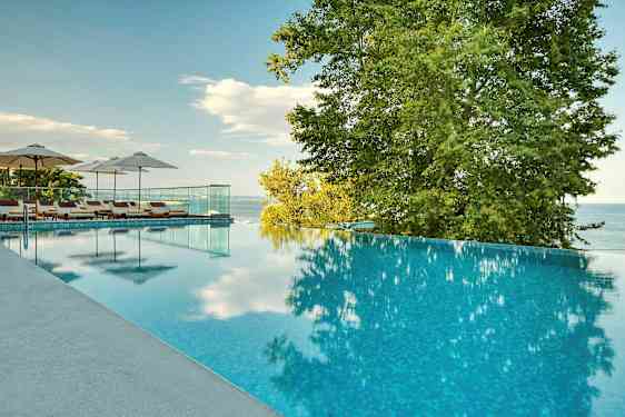 Infinity-allas, jonka vieressä aurinkotuoleja ja sateenvarjoja, ympäröitynä puilla ja näkymällä merelle. (Tekoälyn luoma teksti)