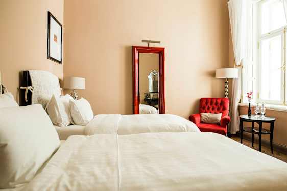 Hotellihuone, jossa kaksi sänkyä, punainen nojatuoli, pieni pöytä ja suuri ikkuna vaaleilla verhoilla. (Tekoälyn luoma teksti)