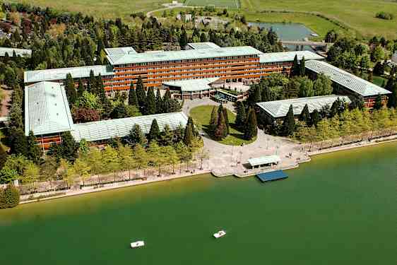 Ilmakuva suuresta rakennuksesta, jonka ympärillä on vihreitä puita ja vesialue, jossa on pieniä veneitä. (Tekoälyn luoma teksti)