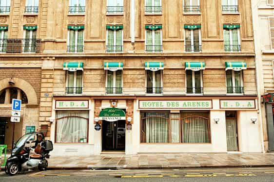 Rakennus hotellijulkisivulla, kyltti tekstillä 'Hotel des Arenes' ja vihreä-valkoiset markiisit ikkunoiden yläpuolella. (Tekoälyn luoma teksti)