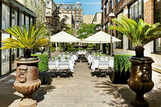 Ulkotila, jossa on pöytiä ja aurinkovarjoja, ympäröitynä rakennuksilla ja palmuilla. (Tekoälyn luoma teksti)