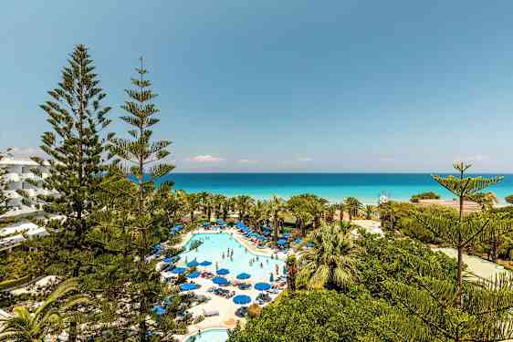 Kuva hotellialueesta, jossa uima-allas ja aurinkotuolit, ympärillä vehreyttä ja näkymä merelle. (Tekoälyn luoma teksti)