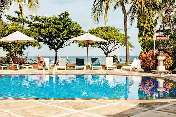 Kuva uima-altaasta, jossa on kirkkaan sininen vesi, ympäröitynä aurinkotuoleilla ja varjoilla, taustalla meri ja palmupuut. (Tekoälyn luoma teksti)