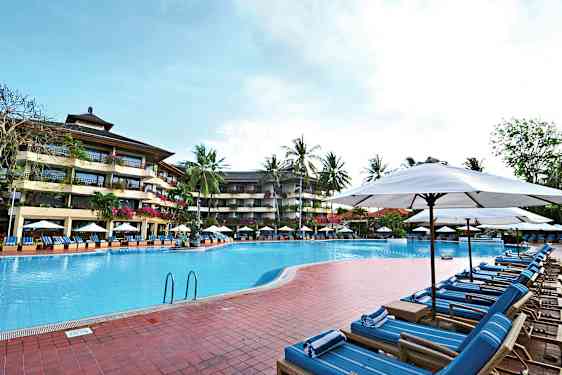 Kuva ulkouima-altaasta, jota ympäröivät aurinkotuolit ja aurinkovarjot, taustalla monikerroksinen hotelli ja palmuja. (Tekoälyn luoma teksti)