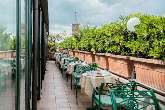 Ulkoterassi, jossa on katetut pöydät ja vihreät metallituolit, reunustettu suurilla viherkasveilla täytetyillä istutuslaatikoilla. (Tekoälyn luoma teksti)