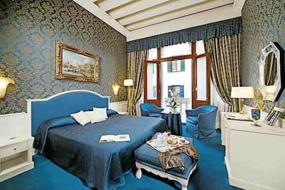 Kauniisti sisustettu hotellihuone, jossa on sinisiä ja kultaisia yksityiskohtia, suuri sänky, oleskelualue ja suuret ikkunat. (Tekoälyn luoma teksti)