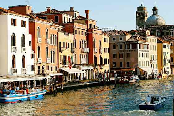 Kuva Venetsian kanavasta, jossa on värikkäitä rakennuksia, veneitä ja kupolimainen kirkko taustalla. (Tekoälyn luoma teksti)