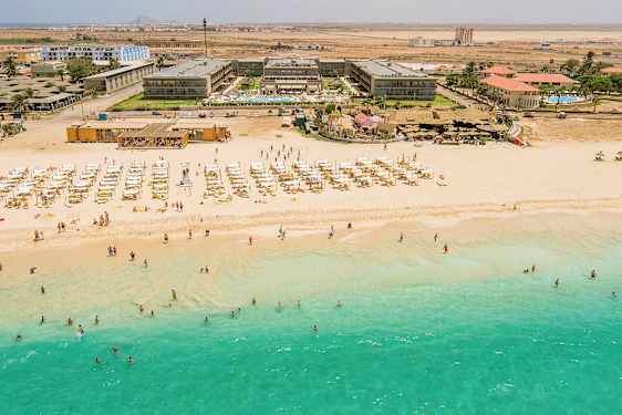 Ilmakuva rannasta, jossa on turkoosia vettä, aurinkotuoleja ja aurinkovarjoja, sekä hotellirakennus taustalla. (Tekoälyn luoma teksti)