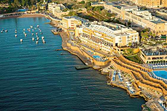 Ilmakuva rannikon hotellista, jossa on uima-altaita ja aurinkotuoleja meren äärellä. Pieniä veneitä on kiinnitetty veteen. (Tekoälyn luoma teksti)