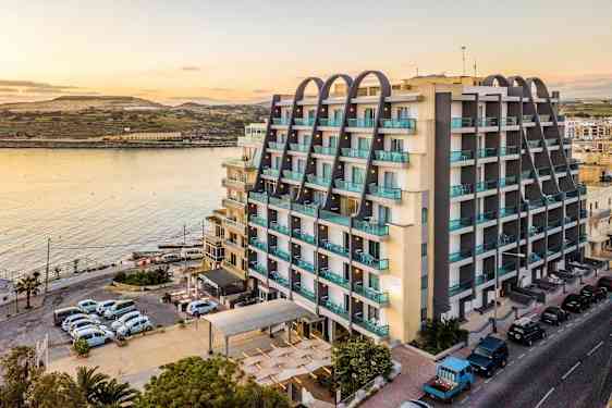 Rakennus veden äärellä, jossa on moderneja kerroksia ja parvekkeita. Etualalla näkyy pysäköityjä autoja ja tie. (Tekoälyn luoma teksti)