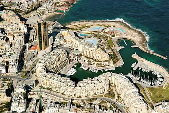 Ilmakuva rannikkokaupungista, jossa on korkeita rakennuksia, venesatama ja taustalla meri. (Tekoälyn luoma teksti)