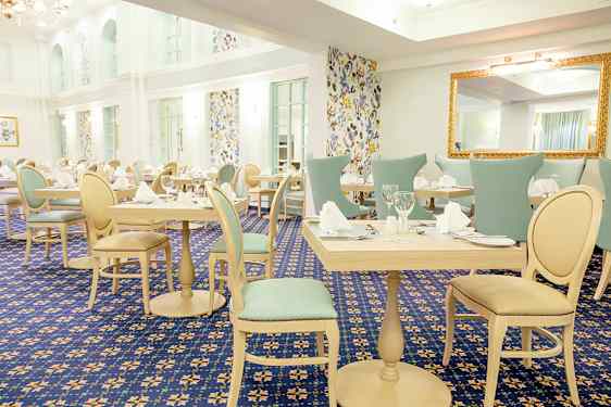 Valoisa ja tyylikkäästi sisustettu ruokailutila, jossa on katetut pöydät ja tuolit, ympäröitynä kuvioiduilla tapeteilla olevilla seinillä. (Tekoälyn luoma teksti)