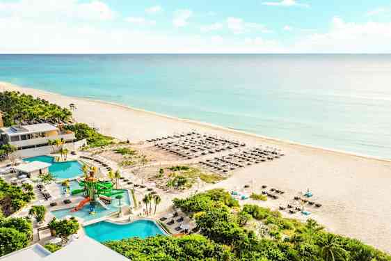 Kuva hiekkarannasta meren äärellä ja sinisen veden ympäröimänä. Rannalla on aurinkotuoleja ja aurinkovarjoja. Etualalla näkyy uima-allas vesiliukumäkien kera sekä ympäröivä vihreä kasvillisuus. (Tekoälyn luoma teksti)