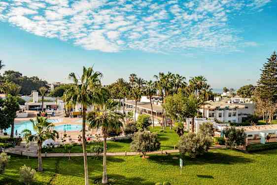 Hotellialue, jossa nurmikkoa, palmuja, uima-allas ja valkoisia rakennuksia. (Tekoälyn luoma teksti)