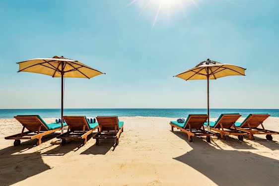 Aurinkotuoleja ja aurinkovarjoja hiekkarannalla, taustalla meri. (Tekoälyn luoma teksti)