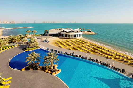 Kuva modernista hotellikompleksista merenrannalla. Etualalla on suuri uima-allas ja rantatuolit. (Tekoälyn luoma teksti)