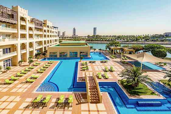 Hotellialue, jossa on suuri uima-allas, aurinkotuoleja ja vihreitä ulkoalueita. Rakennuksessa on useita kerroksia ja se sijaitsee veden äärellä. (Tekoälyn luoma teksti)